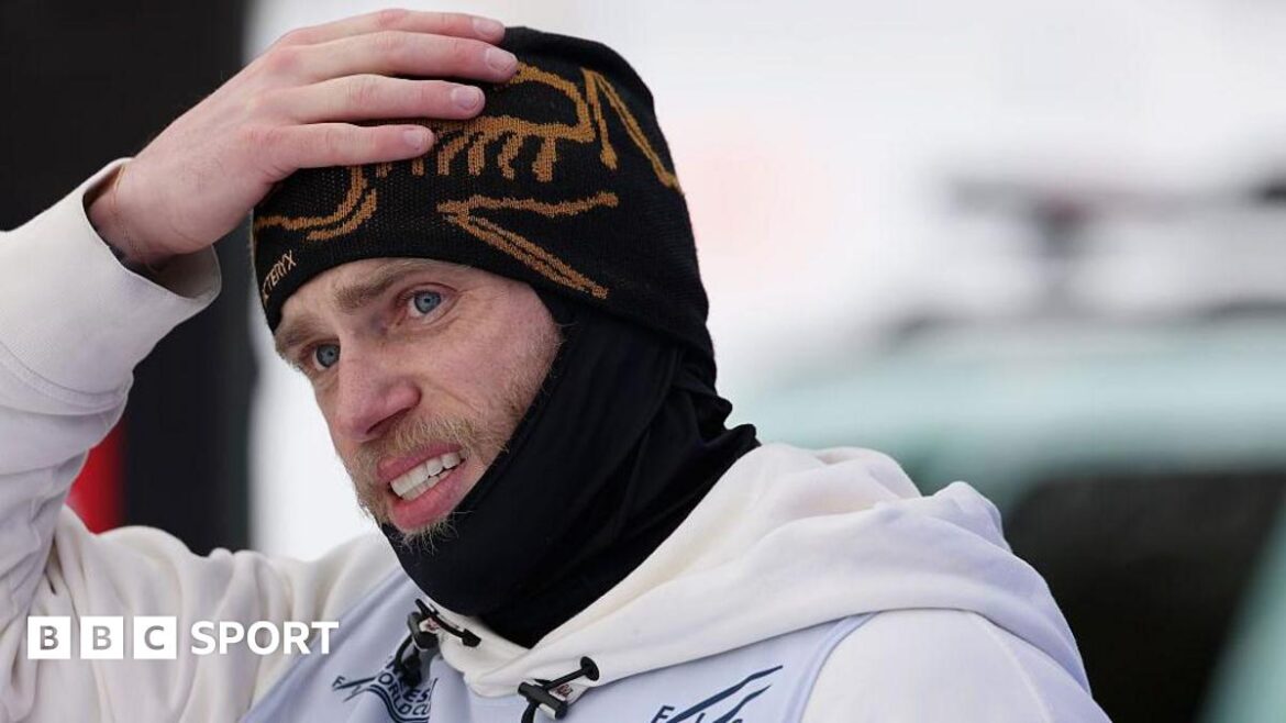 Gus Kenworthy is wearing a black beanie and snood and has his hand on his head