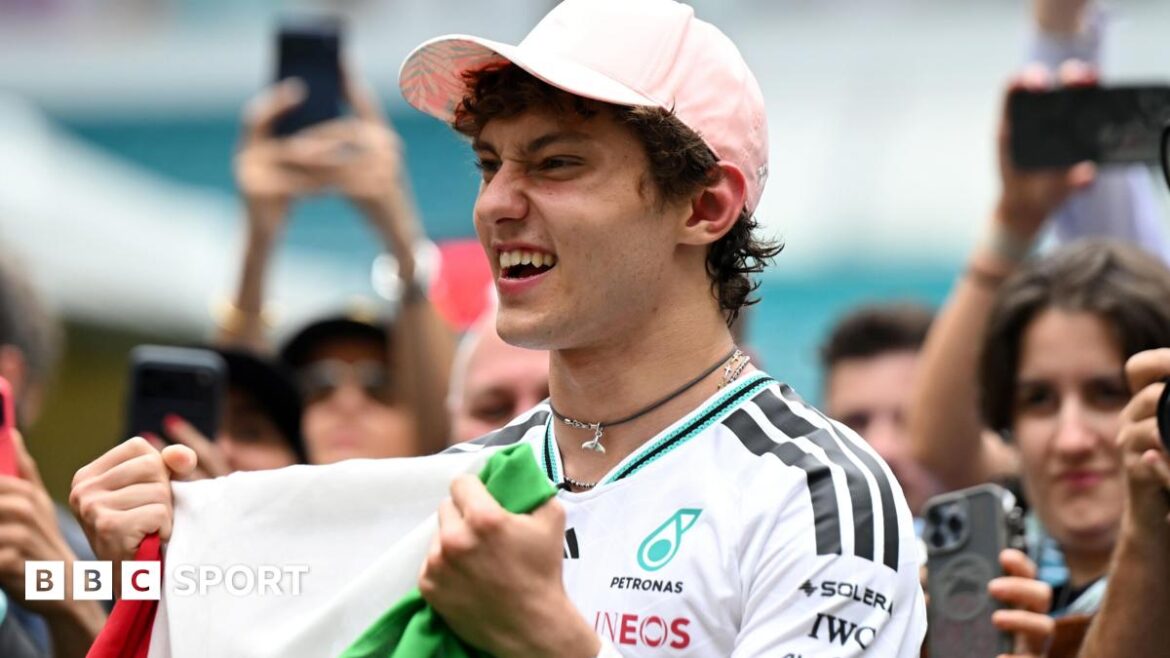 Kimi Antonelli celebrates his race win in Miami while holding the Italian flag. A crowd are gathered around him and taking photographs
